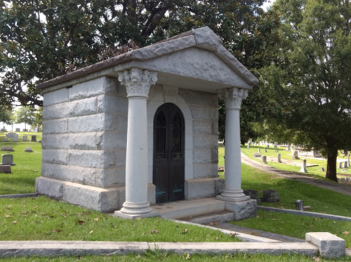 A stone mausoleum with columns and a metal door, surrounded by grass and gravestones in a cemetery.