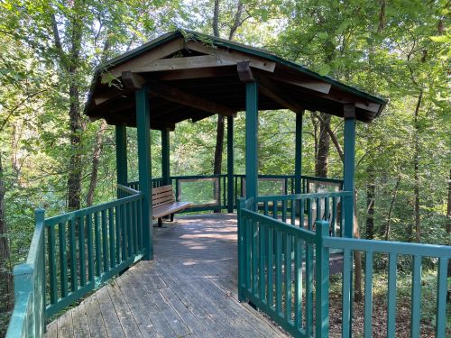 A wooden gazebo with a green railing, surrounded by trees in a forested area.