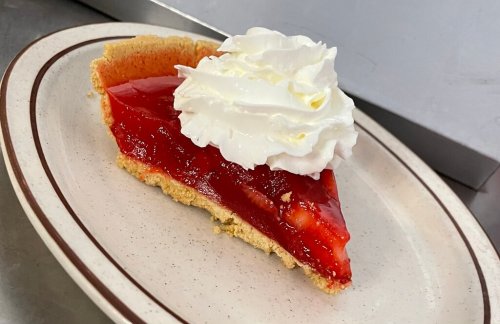 A slice of strawberry pie topped with whipped cream on a white plate with a brown rim.