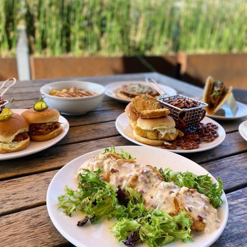 A table with various dishes, including sliders, fried chicken, salad, and sides, set outdoors.