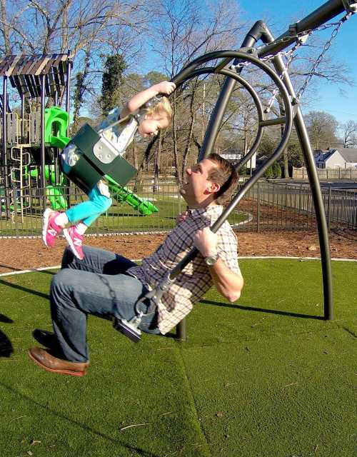 A man swings a young girl on a swing set in a playground, both smiling and enjoying their time together.