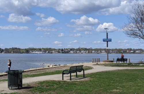 A serene lakeside park with a bench, a person fishing, and a girl walking near the water under a cloudy sky.
