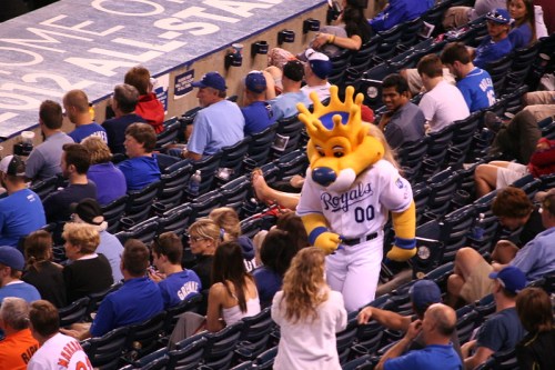 A mascot in a Royals uniform walks through a crowd of fans at a baseball game, engaging with the audience.