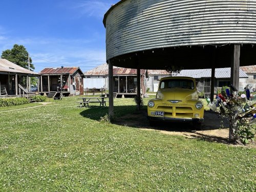 A vintage yellow truck parked under a circular metal structure in a grassy area with rustic buildings in the background.