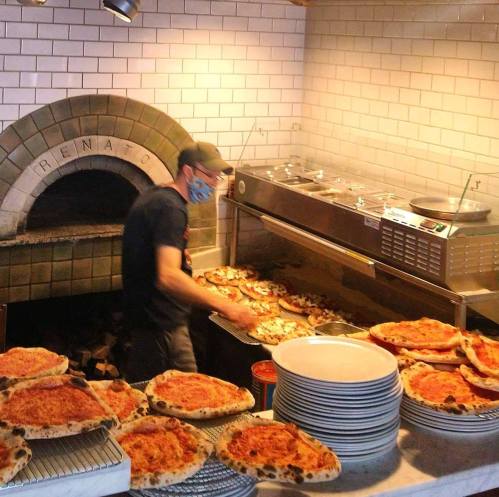 A chef in a mask prepares pizzas in a restaurant kitchen, with freshly baked pizzas stacked on the counter.