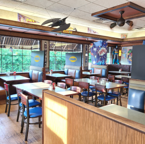 A brightly lit restaurant with Batman-themed decor, featuring empty tables and chairs, and large windows.