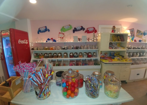 A colorful candy shop display with jars of sweets, lollipops, and a vintage Coca-Cola fridge in the background.