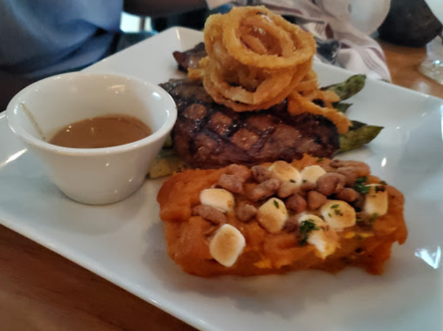 A plate featuring grilled steak, sweet potato topped with nuts, green beans, and a side of sauce. Onion rings on top.
