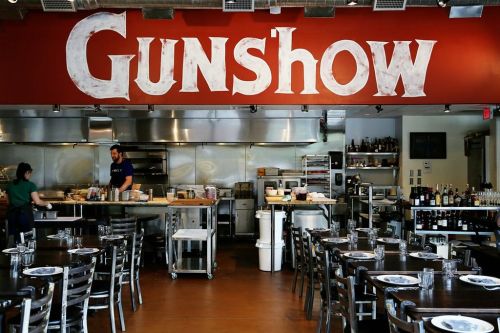 Interior of a restaurant named "Gunshow," featuring a kitchen and dining area with tables set for guests.