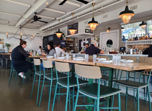 A modern restaurant interior with patrons seated at a long wooden bar, pendant lights, and a stylish decor.