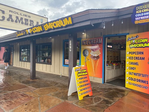 A colorful storefront of "Popcorn Emporium" featuring signs for popcorn, ice cream, and candy on a rainy day.