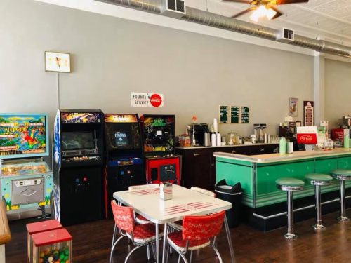 A retro diner interior featuring arcade machines, a green counter, and red stools, with a table in the foreground.