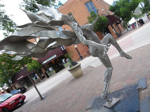 A metallic sculpture of a figure in motion, with flowing elements, set in an urban plaza with shops in the background.