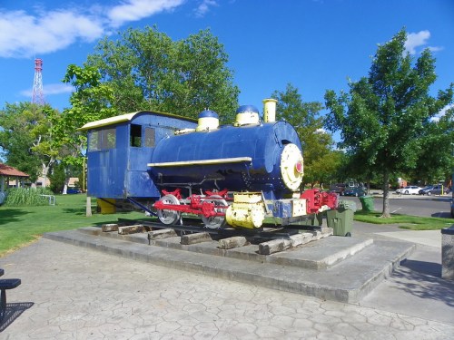 A blue and yellow vintage steam locomotive displayed in a park, surrounded by trees and a clear blue sky.