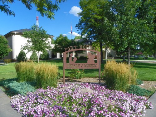 Sign for Fort Mott Library/Museum surrounded by colorful flowers and greenery, with a building in the background.