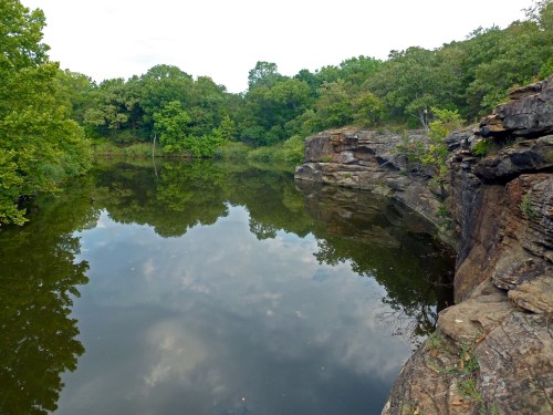 A serene landscape featuring a calm pond reflecting surrounding trees and rocky cliffs under a cloudy sky.