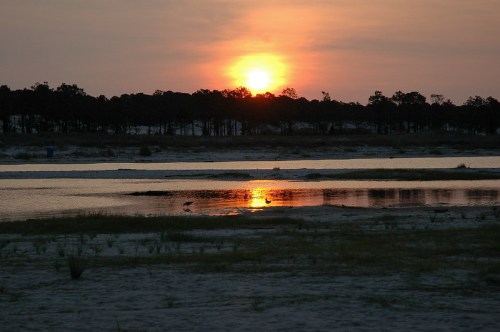 Sunset over a calm river, reflecting orange and pink hues, with silhouettes of trees in the background and sandy shore.
