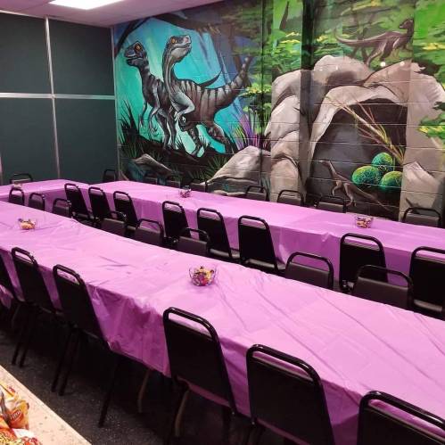 A room with a long table covered in pink cloth, featuring a dinosaur mural on the wall.
