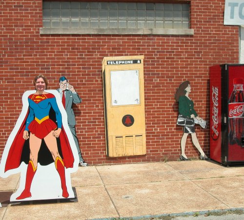 A woman poses with superhero cutouts and vintage telephone booth against a brick wall, with a Coca-Cola vending machine nearby.