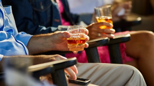 A close-up of hands holding glasses of Maker's Mark whiskey, with people seated in the background.