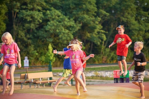 Children in colorful shirts joyfully jumping on a bounce pad at a park, surrounded by trees and outdoor seating.