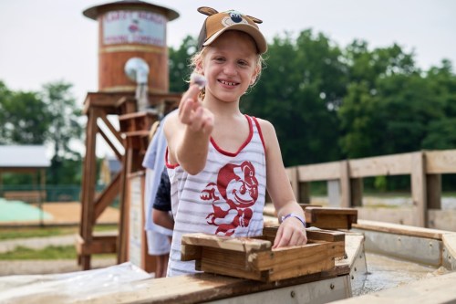 A smiling child in a cap holds up a small stone while sifting through a wooden box outdoors.