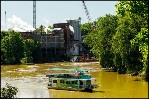 A green boat navigates a muddy river near an industrial area with smoke and cranes in the background.