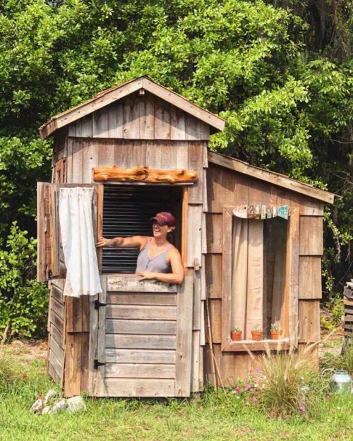 A person stands in the doorway of a rustic wooden shed surrounded by greenery, smiling and holding a curtain.