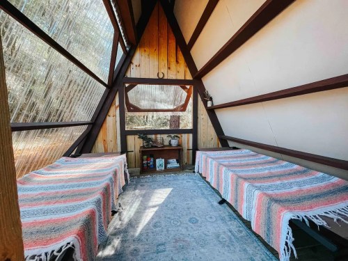 Cozy interior of a cabin with two beds, wooden walls, and a large window overlooking nature.