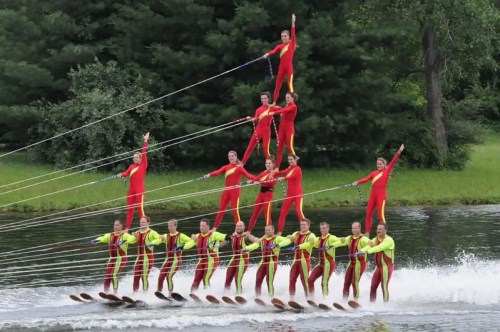 A synchronized water skiing team performs a pyramid formation on a lake, wearing colorful red and yellow suits.