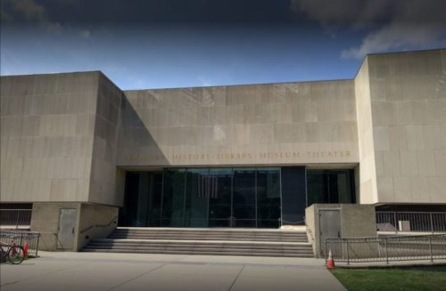 Exterior view of a modern building with large glass doors, labeled "History Library Museum Theater," and steps leading up.