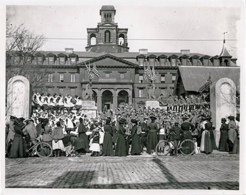 Historic black-and-white photo of a large crowd in front of a grand building, with flags and soldiers present.