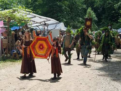 A colorful parade with participants in nature-themed costumes, carrying banners and decorations, walking on a dirt path.