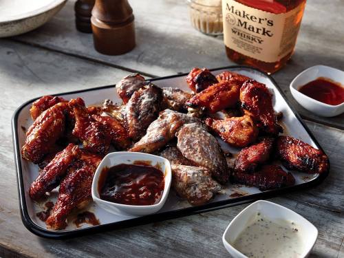 A platter of grilled chicken wings with various sauces, alongside a bottle of Maker's Mark whisky.