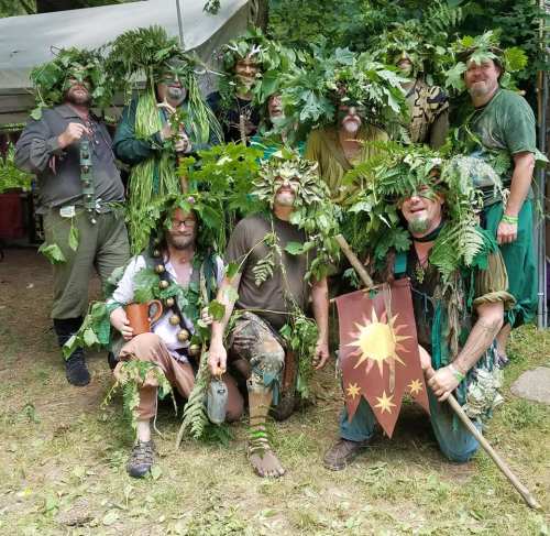A group of men dressed in leafy costumes, posing together in a forest setting, with greenery and props.