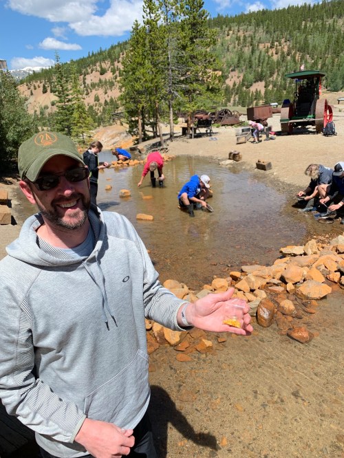 A man smiles while holding a rock near a shallow pond where people are searching for stones. Pine trees and mountains in the background.