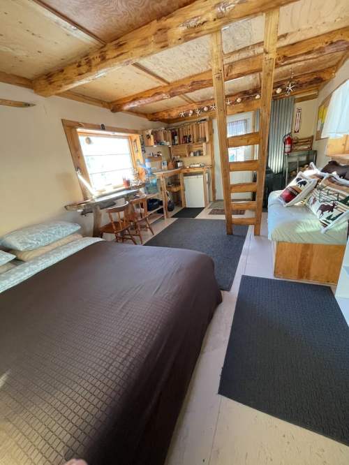 Cozy interior of a small cabin with a bed, seating area, and wooden loft, featuring warm lighting and rustic decor.