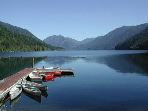 A serene lake surrounded by mountains, with boats docked at a wooden pier under a clear blue sky.