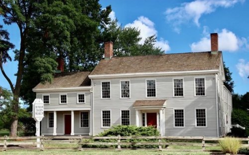 A historic two-story house with white siding, red doors, and a sign, surrounded by trees and a wooden fence.