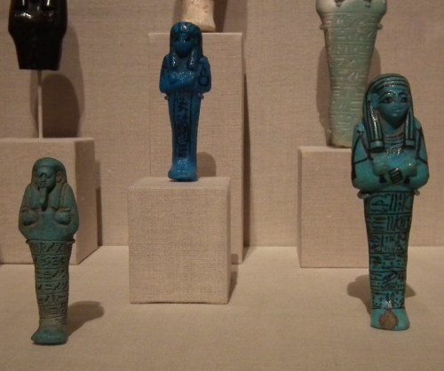 Three ancient Egyptian figurines in turquoise, depicting mummified figures with hieroglyphs, displayed on pedestals.