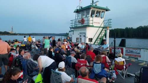 A crowd of people sitting on a dock near a boat, enjoying an outdoor event by the water during sunset.