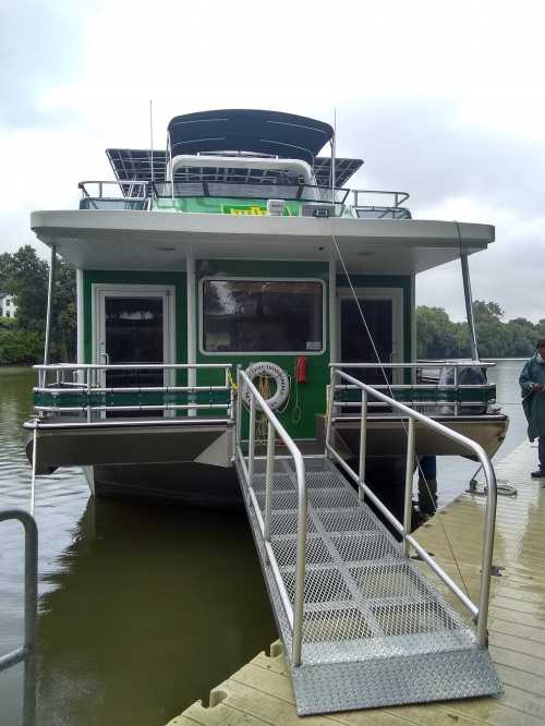 A green and silver boat docked at a pier, with a ramp leading to the entrance and trees in the background.