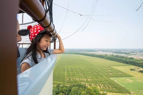 A girl in a Minnie Mouse hat smiles while looking down at a cornfield with "Conner Prairie" carved into it.