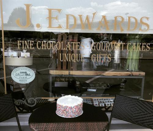 A colorful cake with sprinkles sits on a table outside a shop window displaying gourmet treats and gifts.