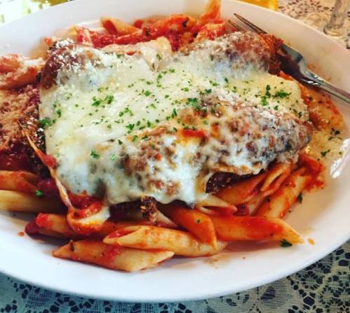 A plate of pasta topped with marinara sauce, melted cheese, and herbs, served with a side of meat.