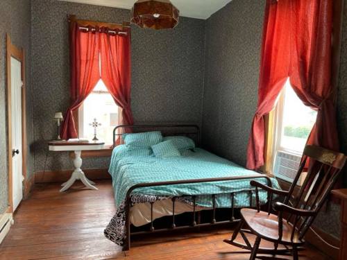 Cozy bedroom with a metal bed, teal bedding, red curtains, wooden floor, and a rocking chair by the window.