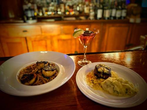 A plate of scallops with noodles, a dish of steak with mashed potatoes, and a cocktail on a bar counter.