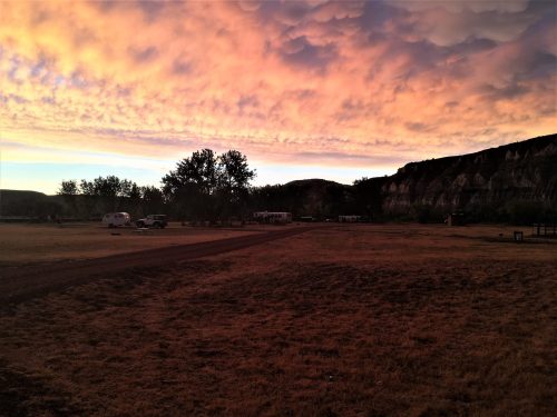 A scenic landscape at sunset, featuring colorful clouds, trees, and a gravel path leading to RVs in the distance.