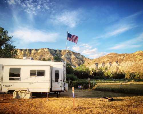 A camper parked in a scenic area with a flagpole displaying the American flag and mountains in the background.