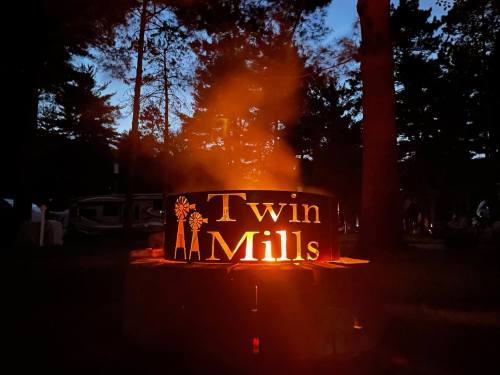 A glowing campfire ring with "Twin Mills" cutout, surrounded by trees at dusk, creating a warm, inviting atmosphere.